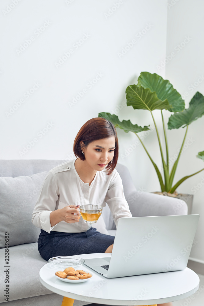 美丽的年轻微笑的亚洲女人在家里的客厅里用笔记本电脑工作，喝咖啡。As