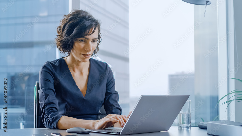 自信的女商人坐在办公桌前，在现代化的办公室里用笔记本电脑工作。时尚吧