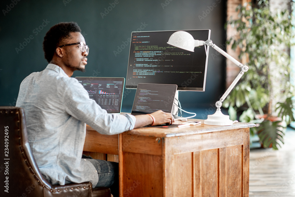 年轻的非洲男性程序员坐在工作场所写程序代码，三个监视器在t中