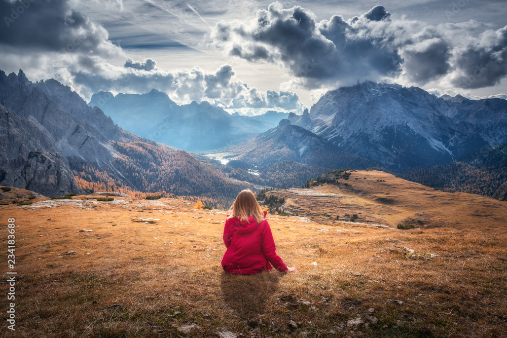 在多洛米蒂，秋天日落时，一位年轻的女士坐在雄伟山脉的山丘上。