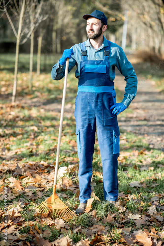 秋天，一名身穿蓝色制服的专业男性清扫工在花园里耙树叶的肖像