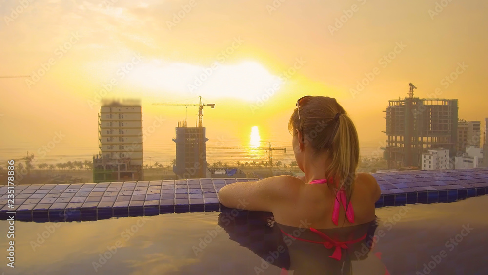特写：身穿粉色比基尼的年轻女子在屋顶游泳池欣赏日落。