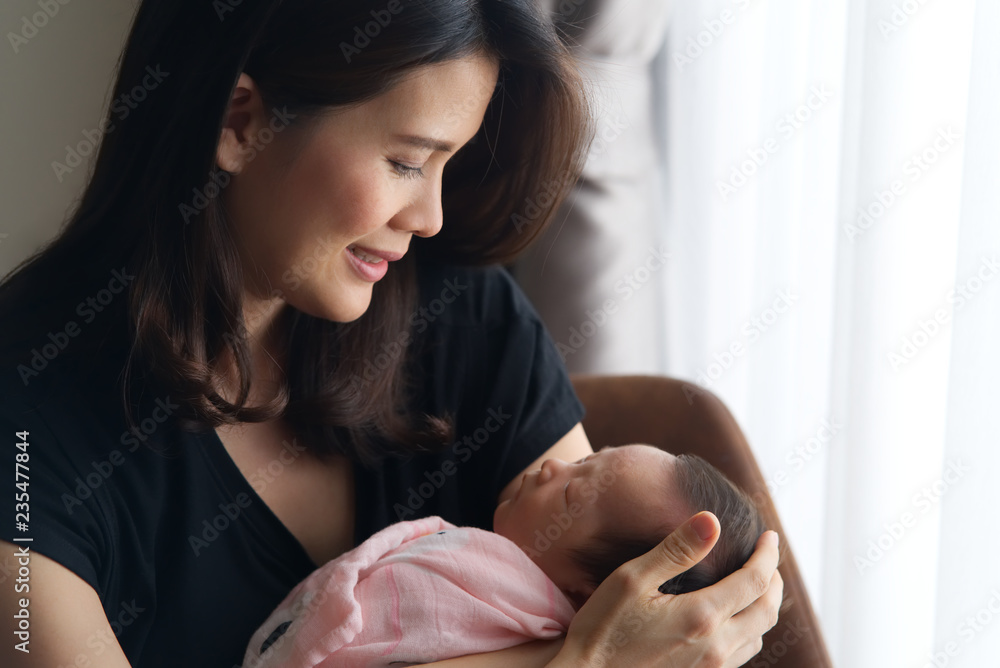 美丽的亚洲微笑母亲抱着刚出生的熟睡婴儿坐在窗户旁边的椅子上。她是
