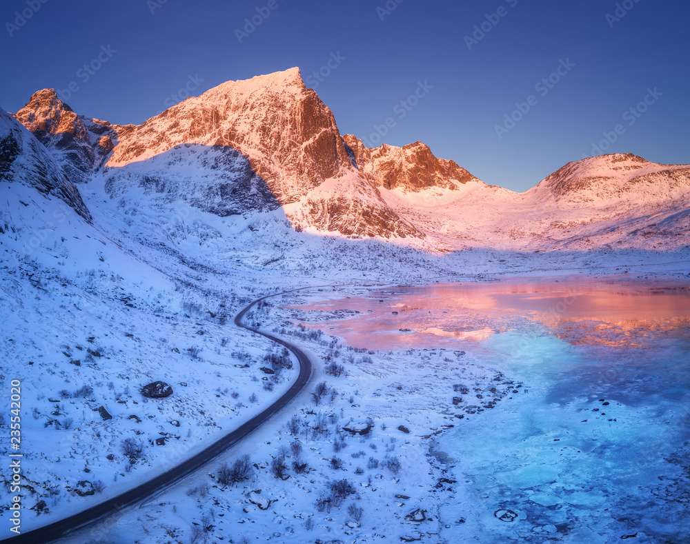 Aerial view of winding road, snow covered mountains and blue sky reflected in water at sunset. Winte