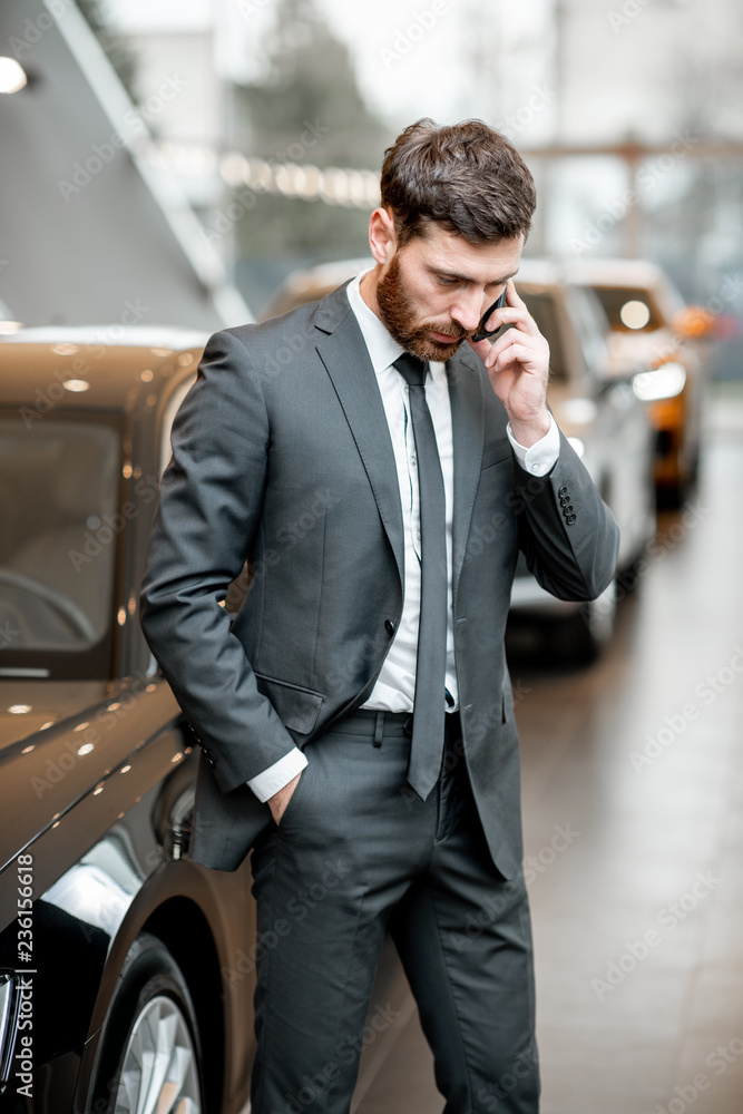 Elegant businessman or salesperson in the suit talking with phone in the car showroom