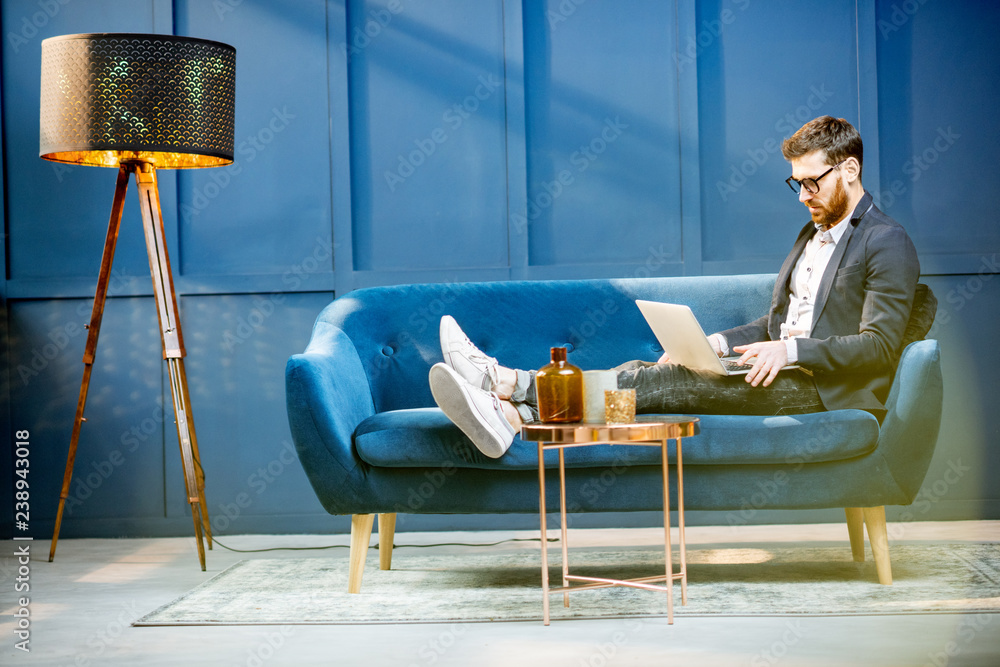 一位优雅的商人的肖像，他拿着笔记本电脑坐在豪华的蓝色办公室的沙发上