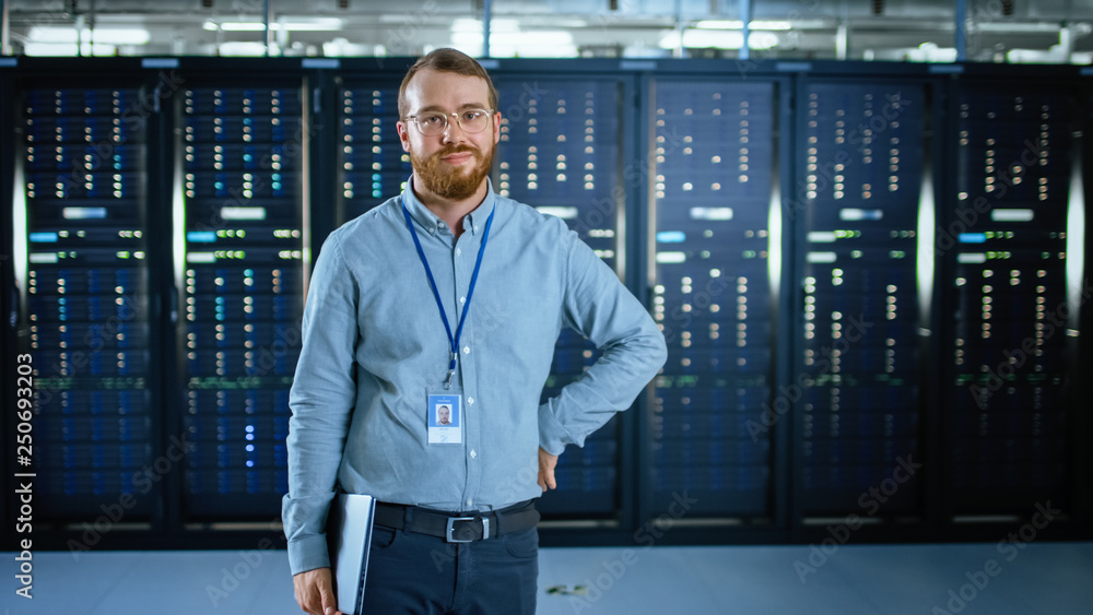 戴着眼镜的IT专家站在数据中心服务器机架旁边，对着摄像头微笑