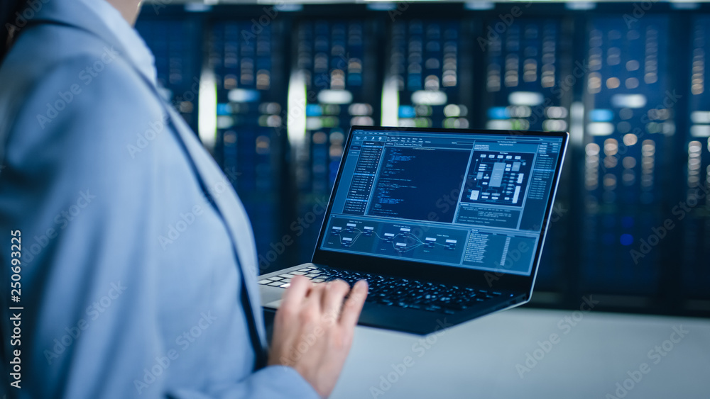 一位IT专家在数据中心服务器机架旁边的笔记本电脑上工作的后视图特写镜头。C