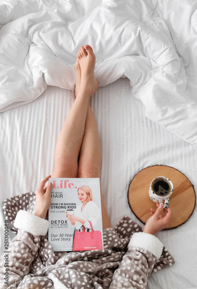 Young woman with fashion magazine drinking coffee on bed in morning