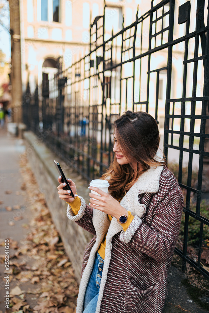 时尚的年轻女子，在城市街道上拿着外卖咖啡和智能手机。