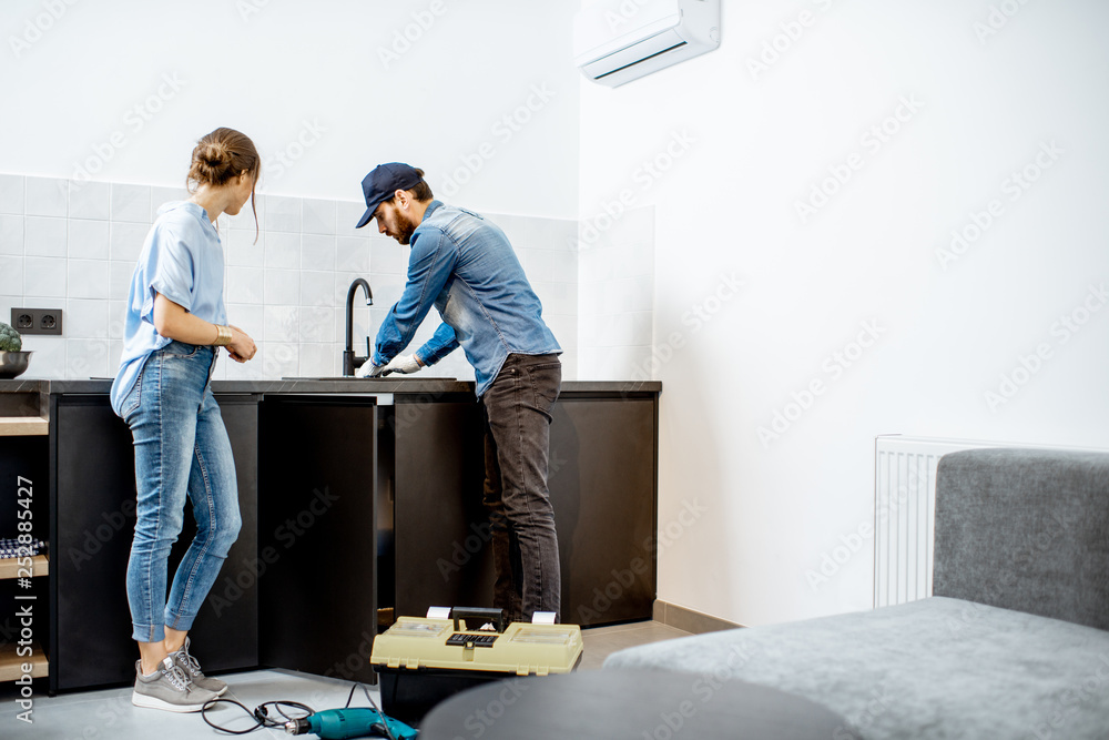 水管工或杂工与年轻女子站在公寓里修理厨房水龙头。家庭维修