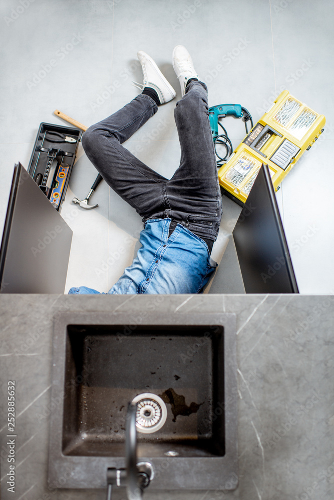 Handyman repairing kitchen plumbing lying under the sink on the floor, view from above