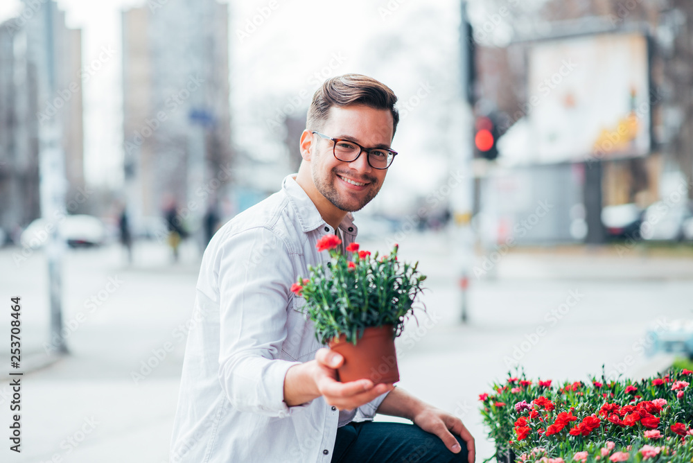 英俊的年轻人在户外花店，手里拿着一盆花，看着相机。