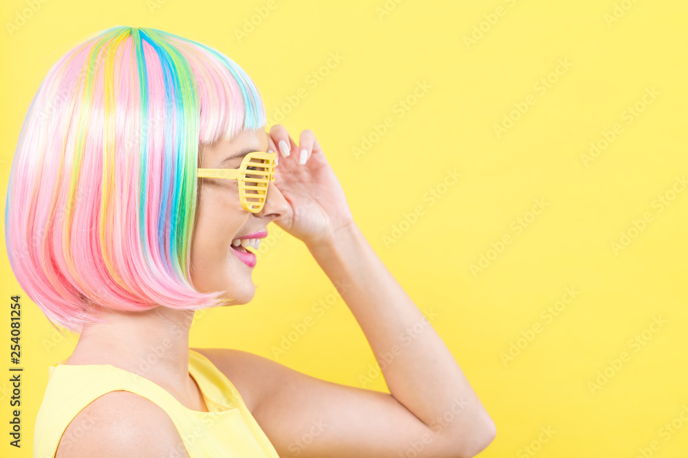 Young woman in a colorful wig with shutter shades sunglasses on a yellow background