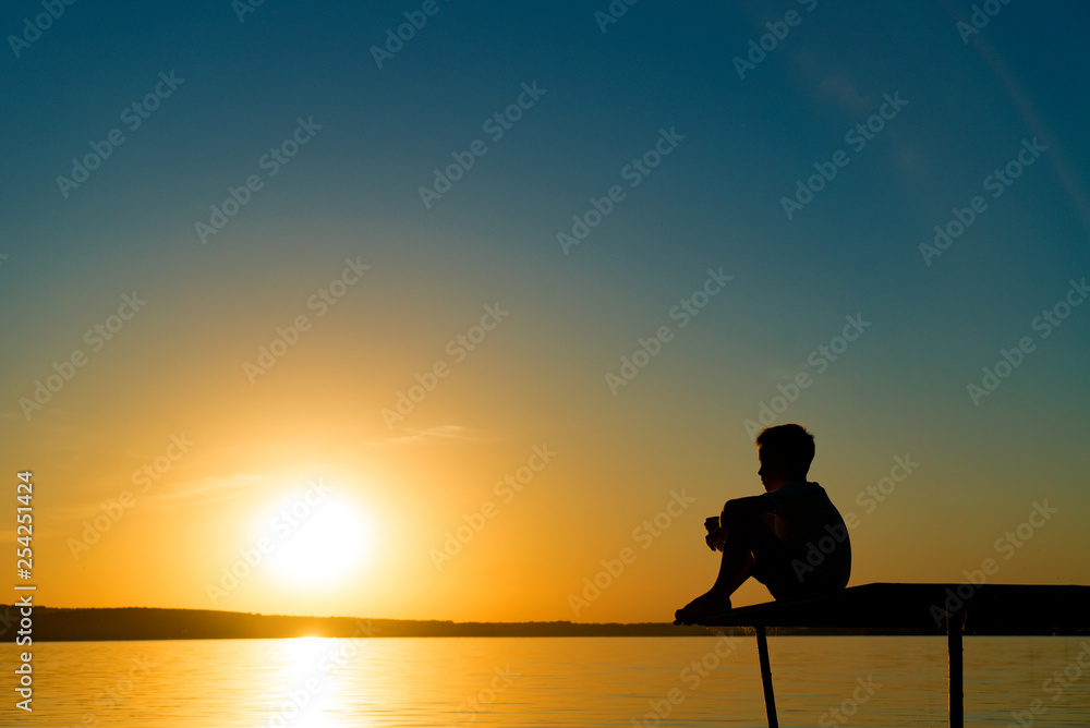 Small boy sitting alone on the footbridge holding his legs and dreaming at sunset. Little kid watche