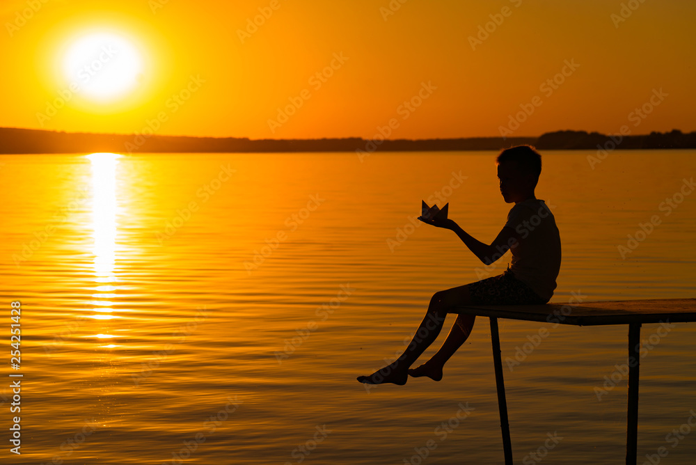 一个小男孩坐在桥上，光着腿拿着折纸船的剪影