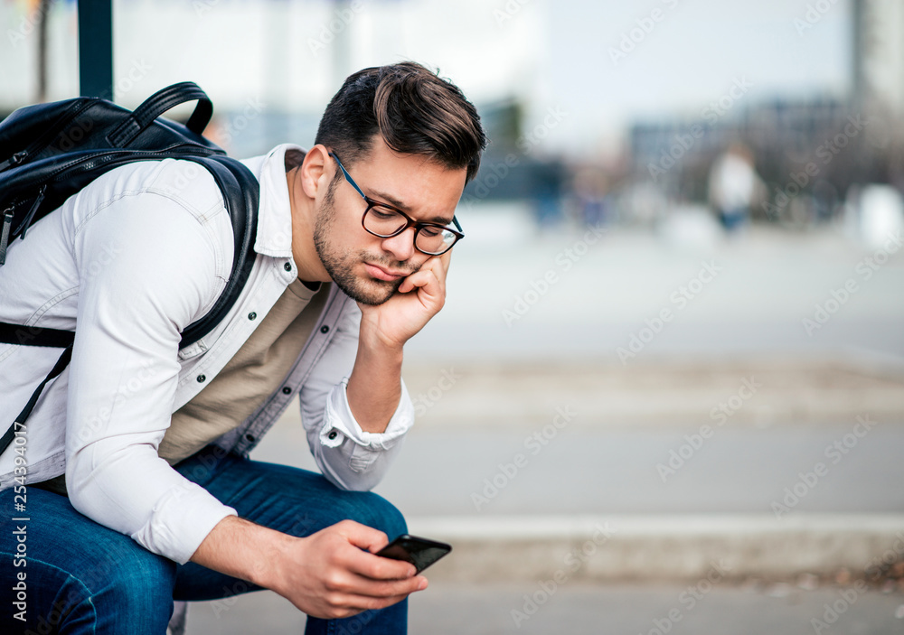 休闲无聊的年轻人背着背包坐在长椅上等公交车，用智能手机。
