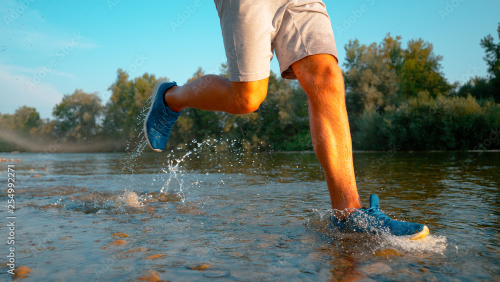 低角度：一名年轻的运动员沿着流经森林的浅河慢跑而下。