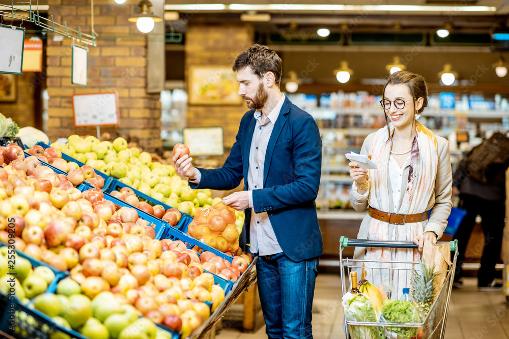 年轻幸福的夫妇站在一起买新鲜水果，购物清单和购物车在超市里