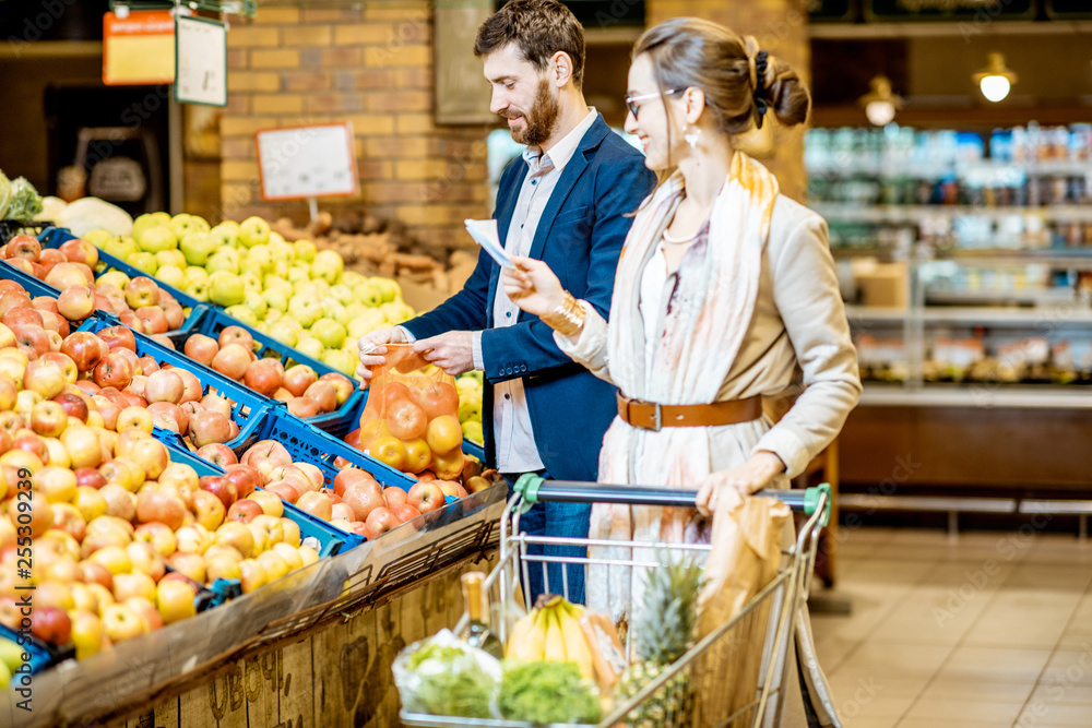 年轻幸福的夫妇站在一起买新鲜水果，购物清单和购物车在超市里