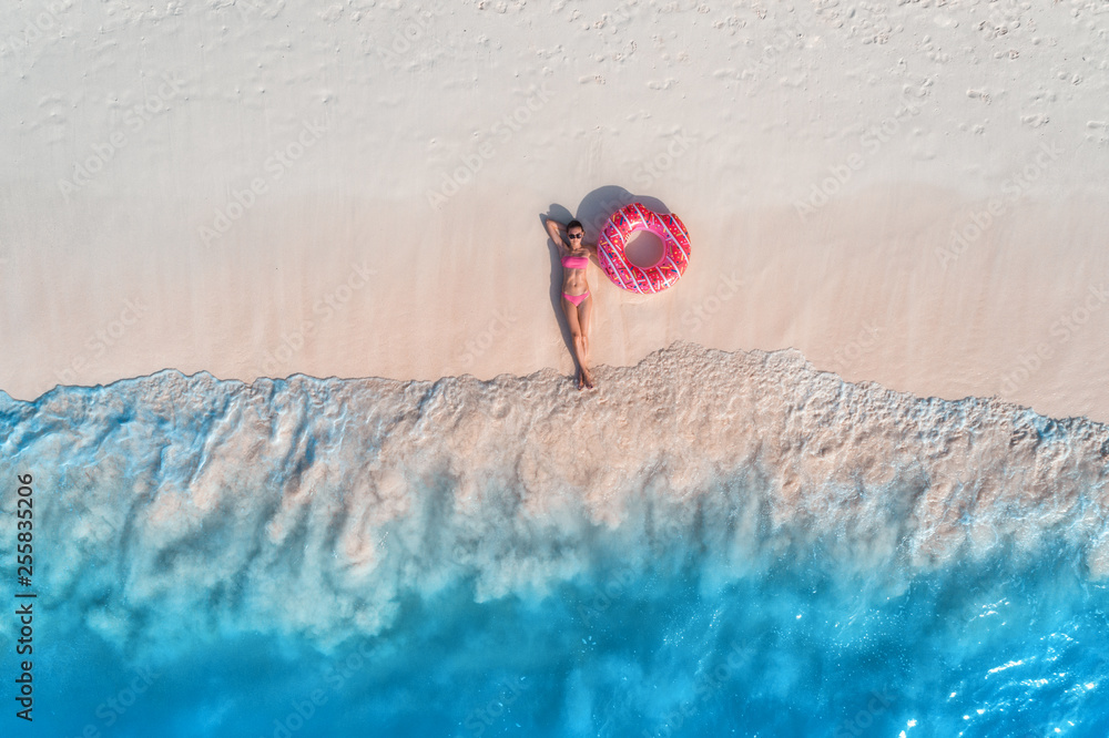 白色沙滩上戴着粉色甜甜圈游泳圈的年轻美女鸟瞰图