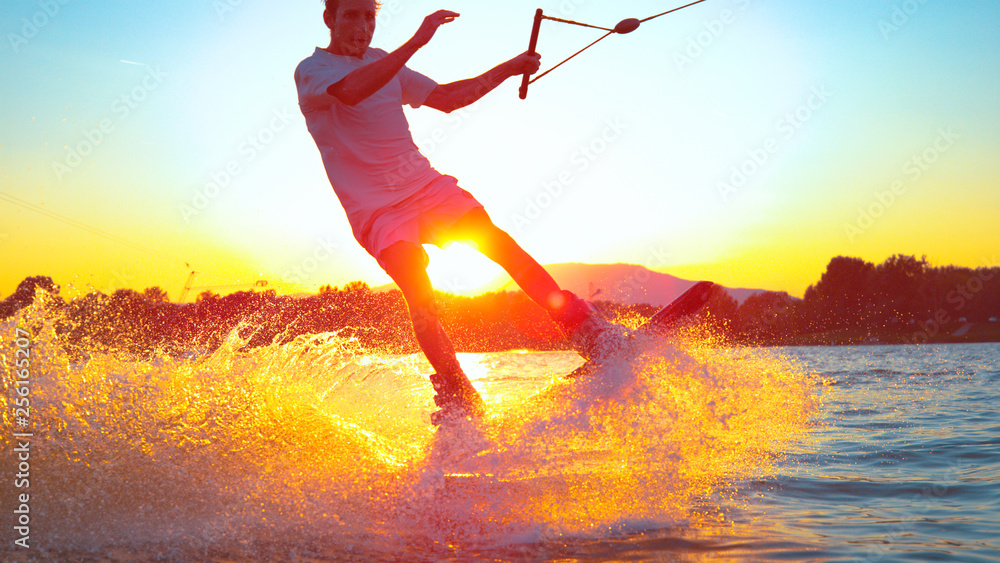 LENS FLARE，CLOSE UP：年轻的冲浪者在日落时滑水并跳180奥利里