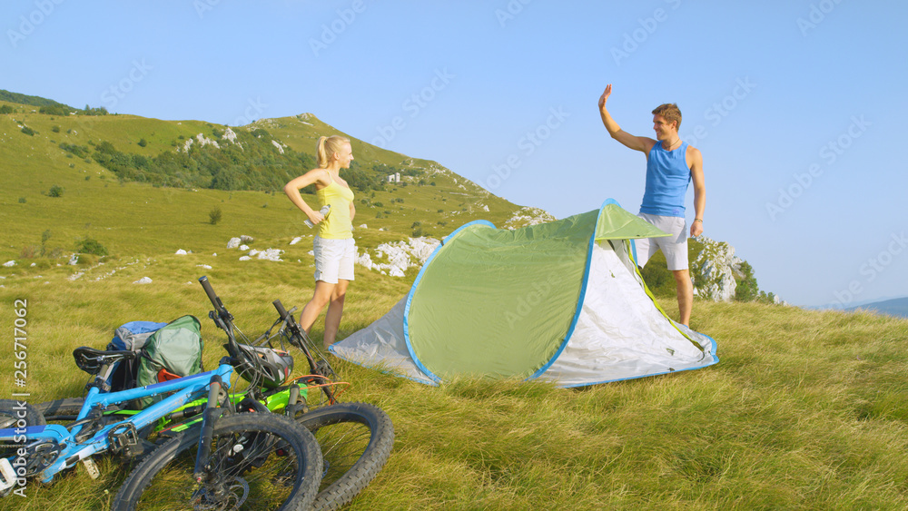 Happy man outstretches hand to high five girlfriend after setting up campsite.
