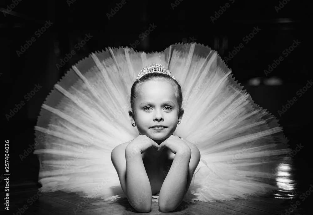 一张黑白照片，照片中一位可爱的微笑芭蕾舞演员穿着白色短裙，头戴王冠，躺在地板上。
