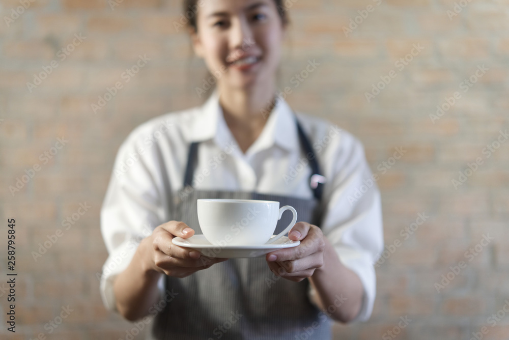 年轻可爱的亚洲咖啡师，穿着漂亮的衬衫，系着围裙，在咖啡馆的白色马克杯里端上热咖啡