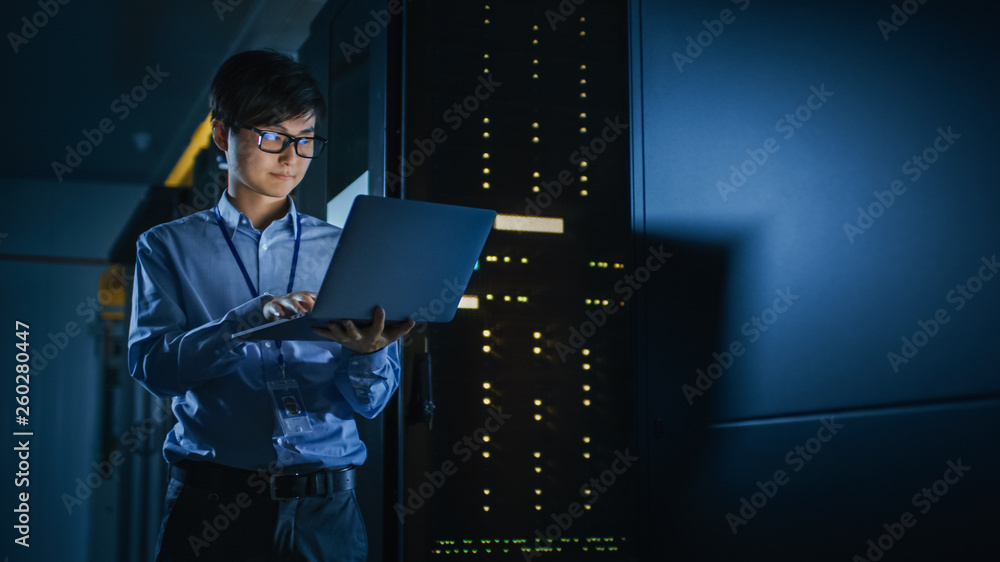 In Dark Data Center: Male IT Specialist Stands Beside the Row of Operational Server Racks, Uses Lapt