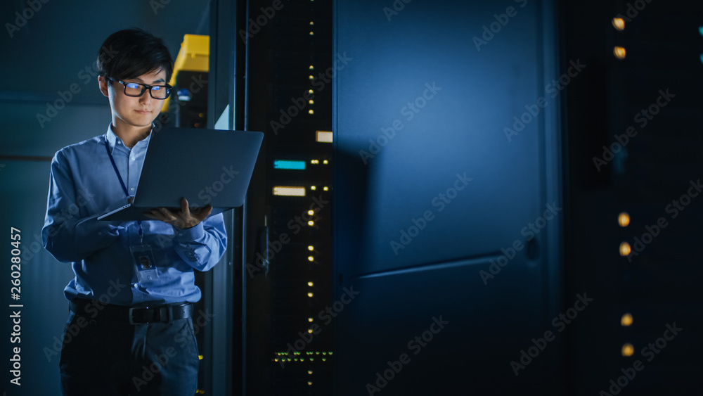 在黑暗的数据中心：男性IT专家站在一排操作服务器机架旁，使用笔记本电脑