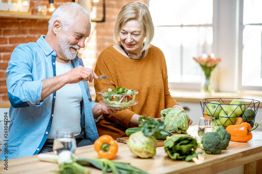 Cheerful senior couple eating salad standing together with healthy food on the kitchen at home. Conc