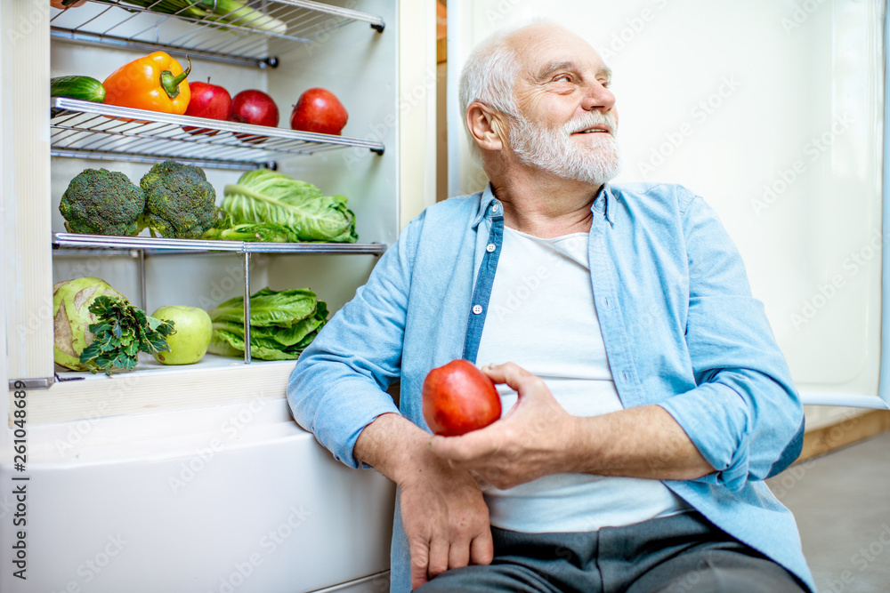 一位体贴的老人坐在装满健康食品的冰箱旁，手里拿着苹果
