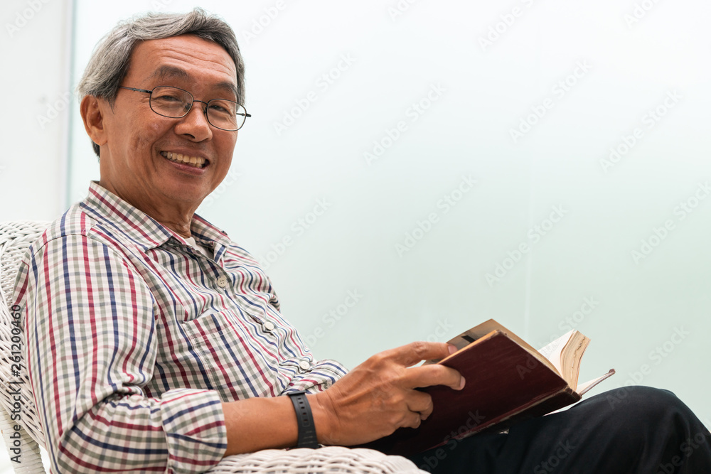 Happy senior Asian man reading book on the chair in living room at home. Retirement lifestyle and ag