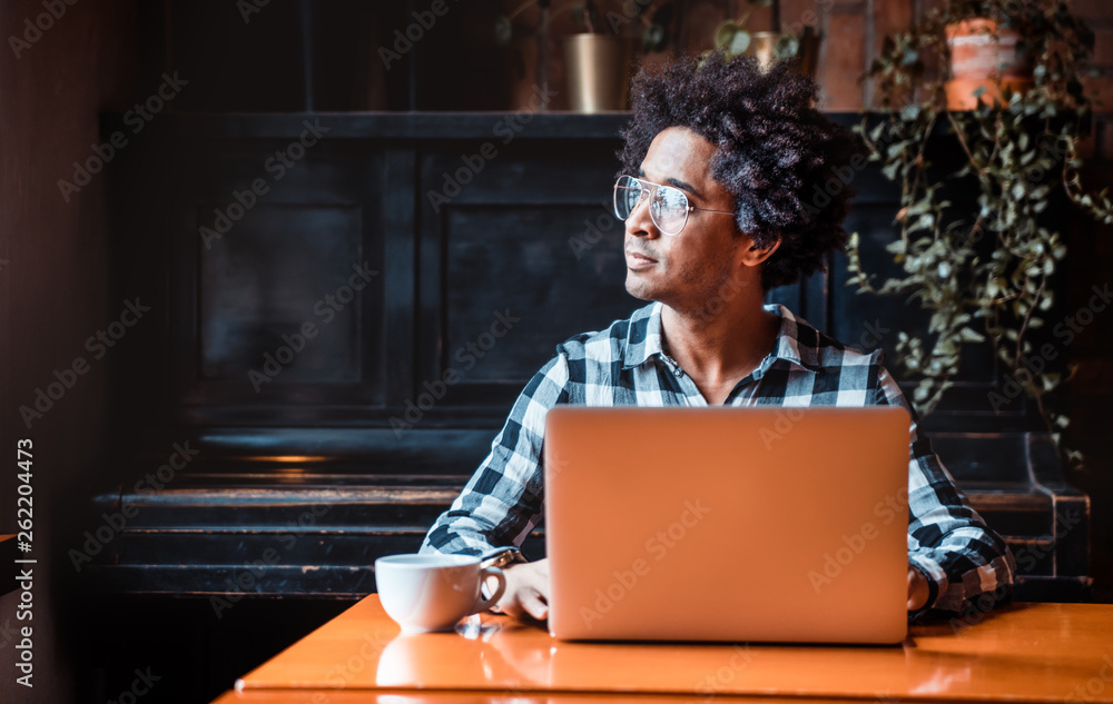 戴眼镜的非洲男子坐在餐厅里用笔记本电脑，年轻人工作的概念