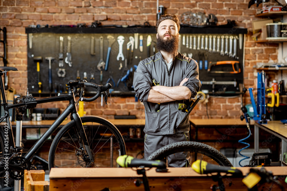 一个英俊的留胡子的修理工穿着工作服，拿着扳手站在自行车车间的肖像