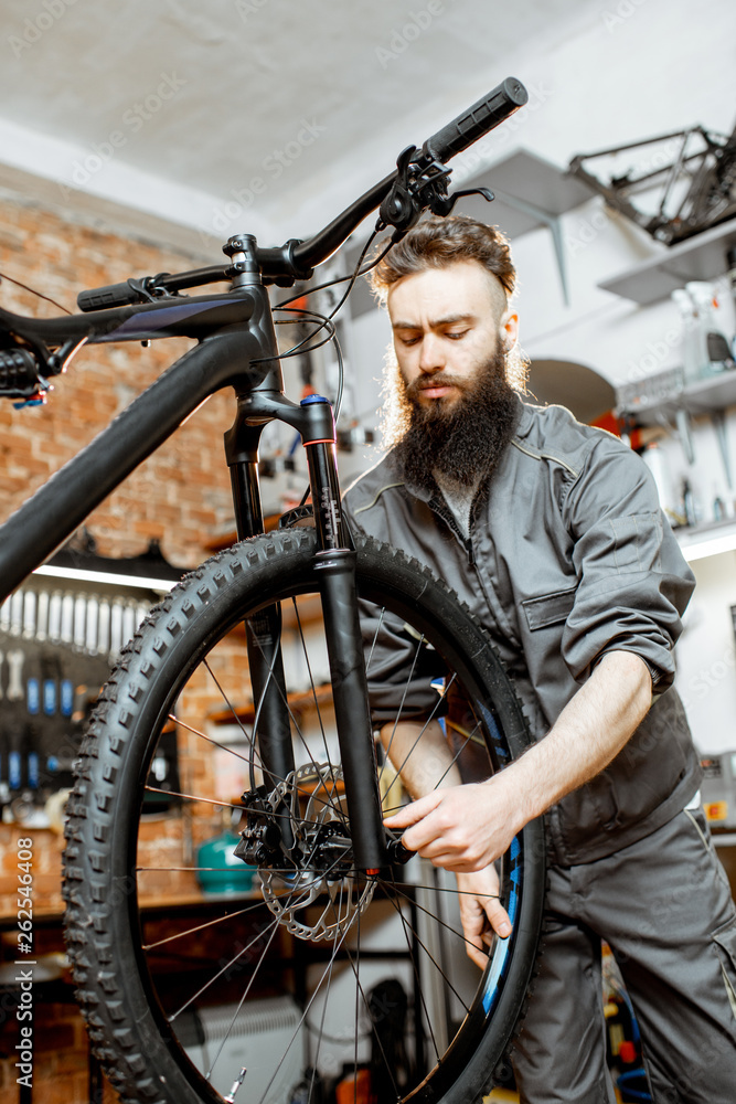 英俊的大胡子修理工穿着工作服在车间的山地自行车上安装轮子