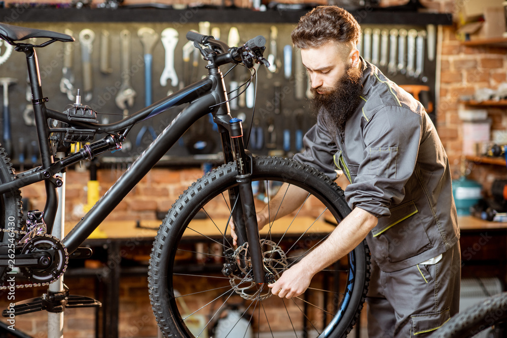 英俊的大胡子修理工穿着工作服在车间的山地自行车上安装车轮