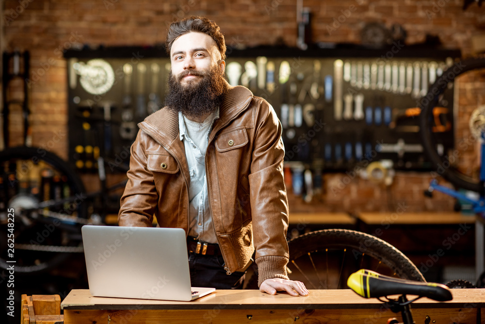 Portrait of a handsome bearded man as bicycle store owner or manager standing with laptop at the bic