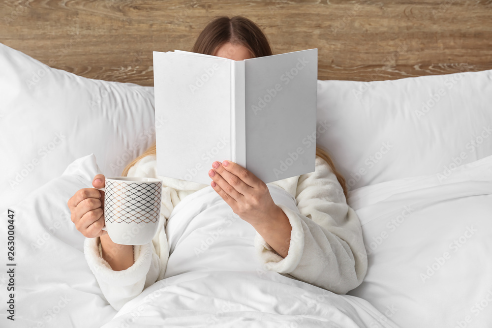 年轻女子在床上拿着一杯热饮看书