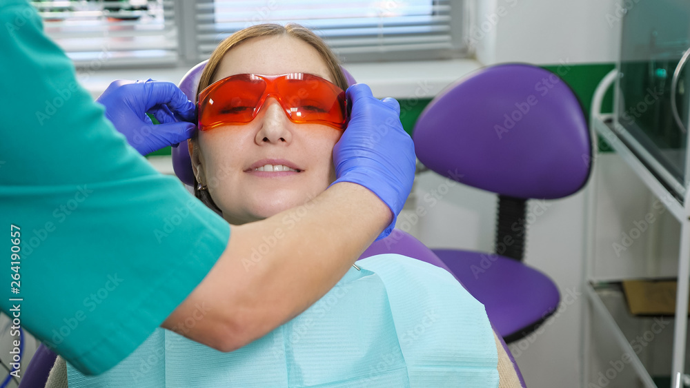 牙医在检查前用手给坐在牙科椅上的年轻女性戴上眼镜。