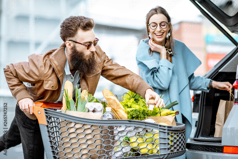 Young stylish couple with shopping cart full of fresh food, packing products into the car on the out
