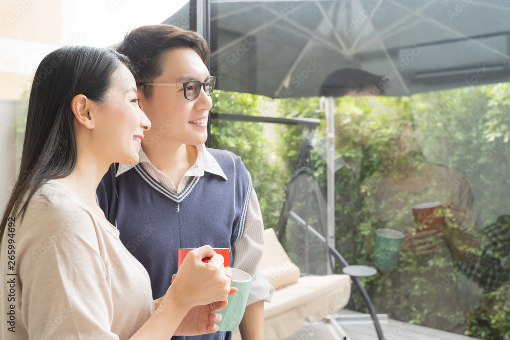 一对年轻的亚洲夫妇在现代化的家里喝咖啡放松身心，室内眺望室外