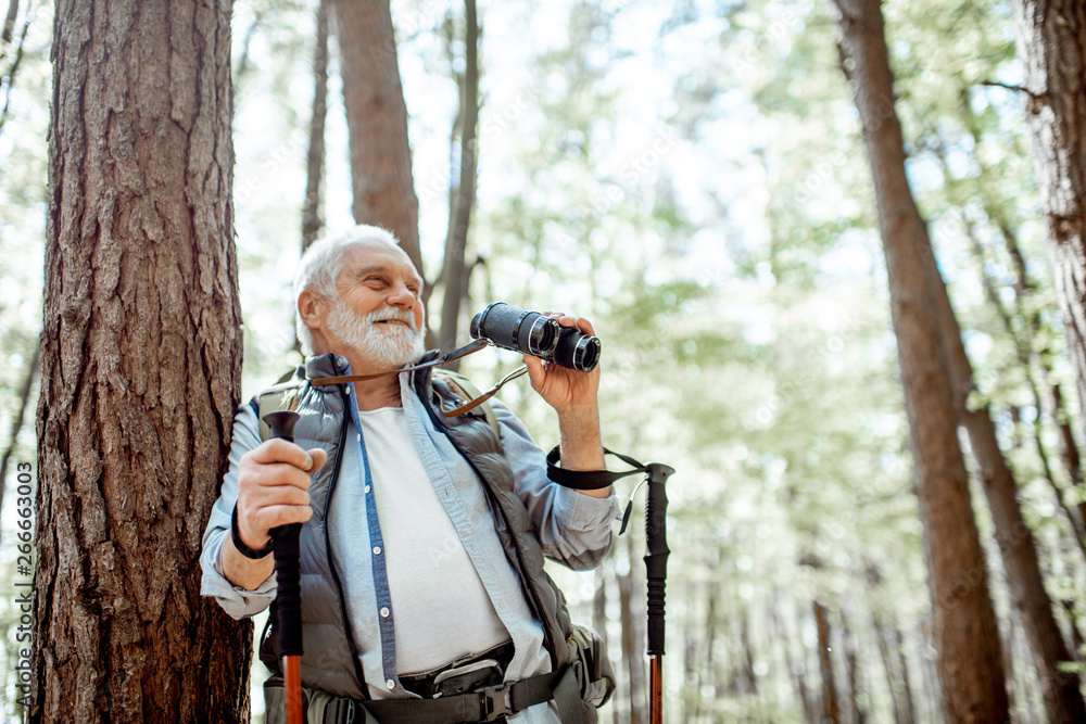 Portrait of a senior man with binoculars and backpack resting near the tree while traveling in the f