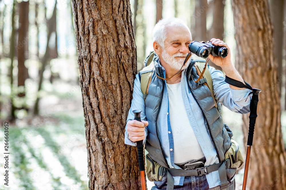Portrait of a senior man with binoculars and backpack resting near the tree while traveling in the f
