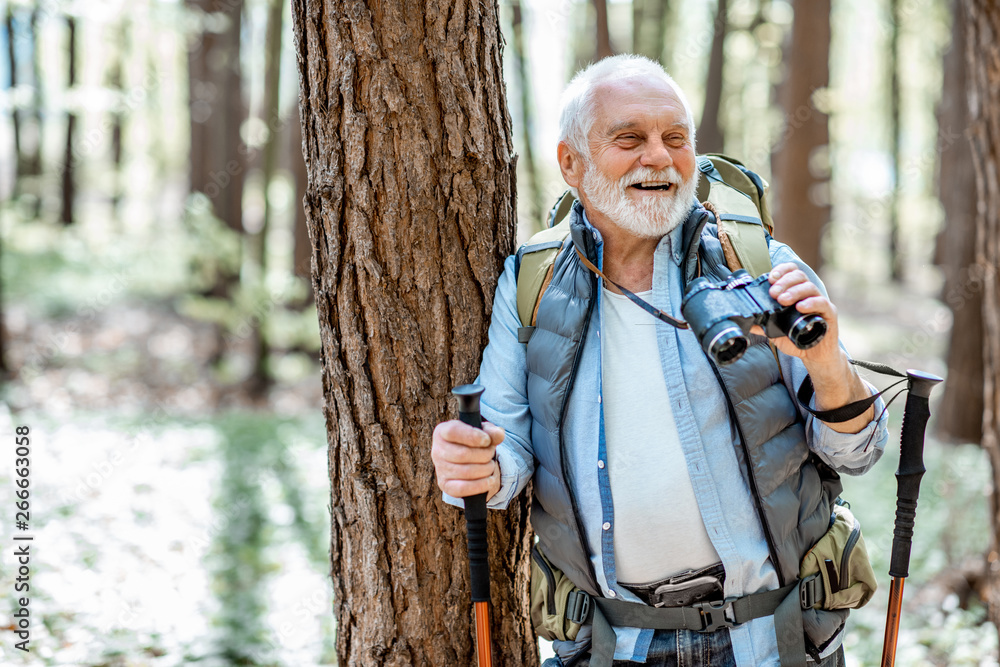 一名老年男子的肖像，他带着双筒望远镜和背包，在森林中旅行时停在树附近