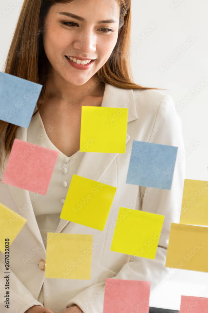 快乐的女商人在办公室的玻璃墙上贴着便签，思考创意。工作计划