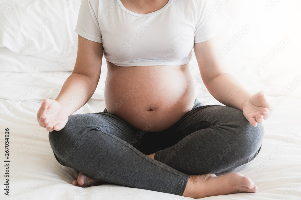 Pregnant woman doing yoga exercise on bed in bedroom at home while taking care of her child. The hap