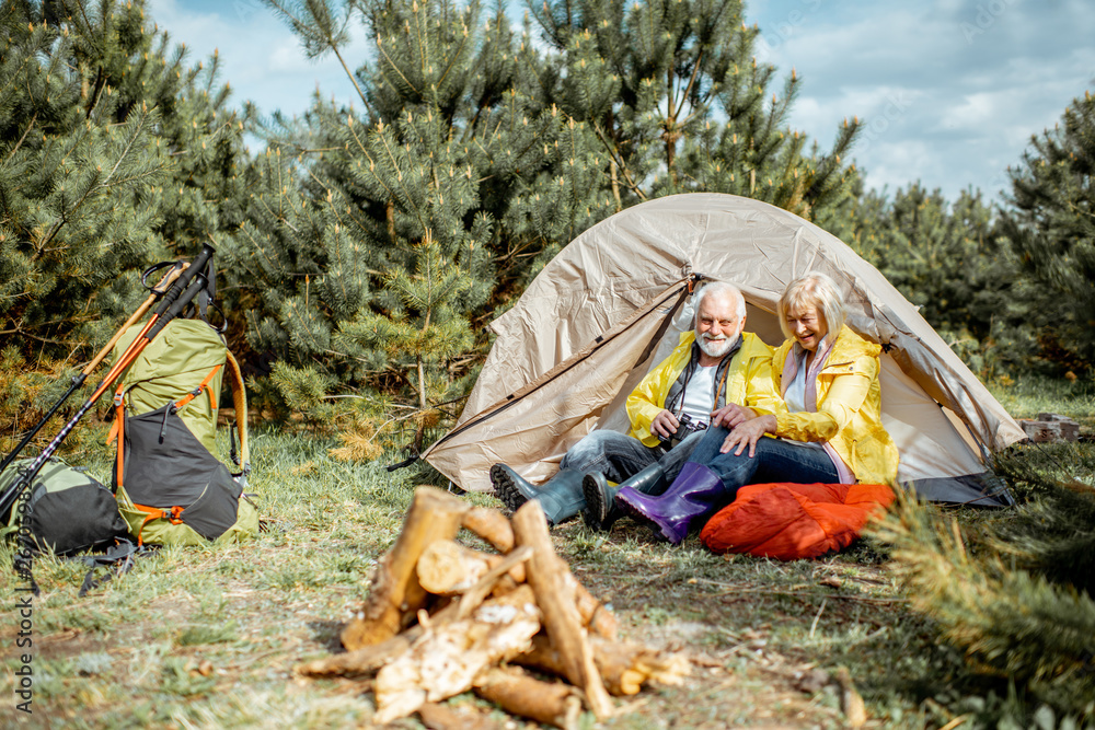 一对穿着黄色雨衣的老年夫妇坐在露营地的帐篷附近，帐篷里有壁炉。