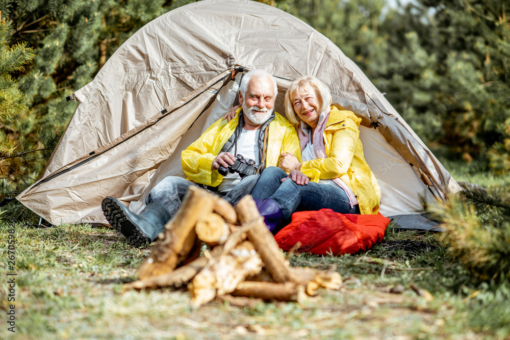 Senior couple in yellow raincoats sitting together near the tent at the campsite with fireplace in t